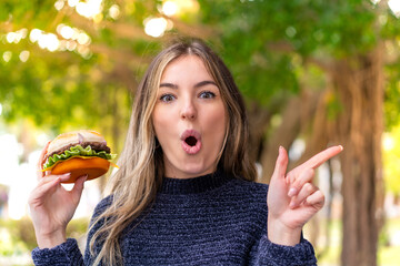 Young pretty Romanian woman holding a burger at outdoors surprised and pointing side