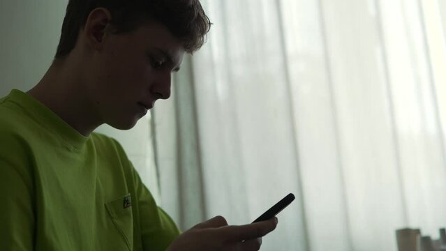 silhouette of a teenager browsing a smartphone while sitting on the couch at home Young curly handsome boy male student chatting texting typing and using mobile phone indoors Caucasian man uses cel