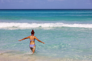 Tanned girl in swimsuit going to swim in blue sea water with her arms outstretched. Beach vacation on Caribbean islands