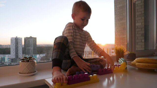 Small independent child plays children constructor at table by window at sunset shine. Inventive preschoolers funny boy collects crafts from details. Kids educational games. Dream of happy childhood