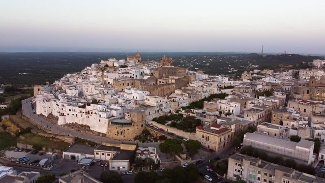 Aerial sunset panoramic drone footage of Ostuni - La Citta Bianca (white city), Puglia, Italy. View of walls, Cathedral, Porta Nova, medieval historic old town landmark tourist destination from above.