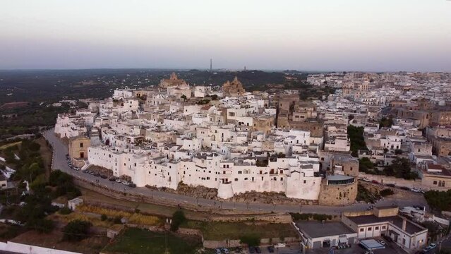 Aerial panoramic drone footage of Ostuni - La Citta Bianca (white city), Puglia, Italy at sunset. View of Cathedral, Porta Nova, the medieval historic old town landmark tourist destination from above.