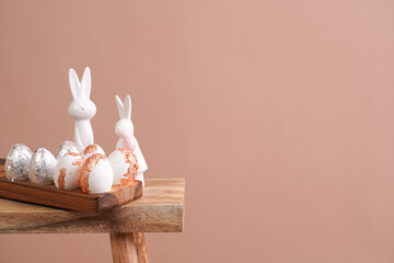 Easter decoration: eggs with silver and rose-gold metallic foil in a bamboos form on a wooden mediterranean style stool, white easter bunny figurines on beige colored seamless background, copy space