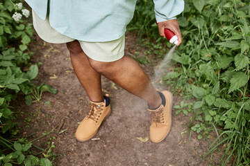 High angle closeup of black woman spaying legs with bug repellent while travelling in nature 