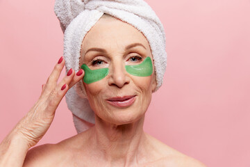 Close-up of beautiful middle-aged woman with towel on head posing with under eye patches, posing on pink studio background. Concept of natural beauty, face skin care, cosmetology and cosmetics, health