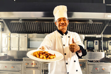 Professional senior chef wearing asian chef's hat in modern hotel kitchen showing delicious Thai food menu ready to serve customer thumbs up happy cooking.