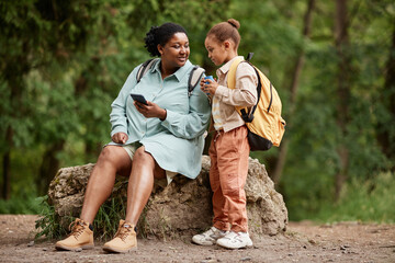 Full length portrait of black mother and daughter sitting on rock while hiking together in nature,...