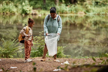Full length portrait of eco-activist mother and daughter cleaning urban trash by river, copy space  - obrazy, fototapety, plakaty