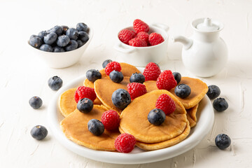 Top view of pancakes on plate with blueberries and raspberries with bowls and jar on white table, selective focus, horizontal