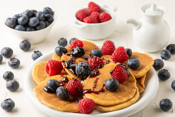 Top view of pancakes with caramel on plate with blueberries and raspberries with bowls and jar on white table, selective focus, horizontal