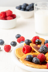 Close-up of plate of caramel pancakes with blueberries and raspberries with bowls and glass of milk, on white table, selective focus, vertical