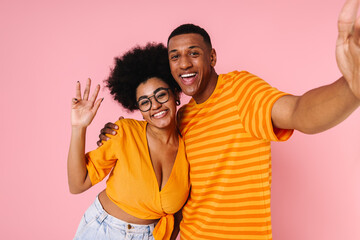 Fototapeta premium Cheery african couple taking selfie while standing isolated over pink background