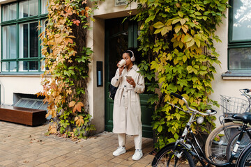 African girl listening music with headphones and drinking coffee while standing outdoors