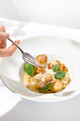 Woman hand holding fork with sea scallop over creamy risotto. Person eat seafood risotto with scallops. Female hand with italian risotto. Trendy menu concept.