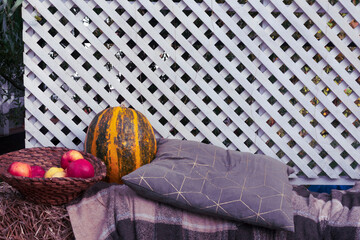 Decorative composition of a pumpkin, apples in a basket, pillow, and knitted plaid with a white wooden background in autumn.
