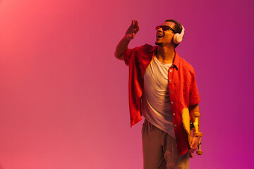 Excited african man holding skateboard and dancing isolated over multicolored neon background