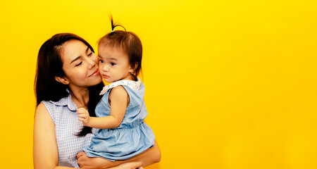 Mother's happiness : Beautiful young mother stands in her studio yellow room holding her cute youngest daughter lovingly kissing the cheek, caring for the joyful innocence of a toddler girl.
