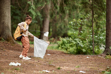 Side view portrait of eco activist little girl picking up plastic bottles in nature trail, copy...