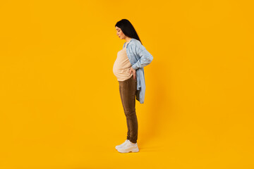 Fototapeta na wymiar Maternity concept. Pregnant woman touching back while standing over yellow background, side view, full length shot