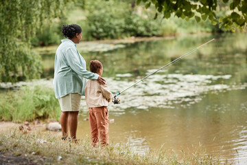Back view of mother and daughter fishing together by lake in beautiful family bonding moment, copy...