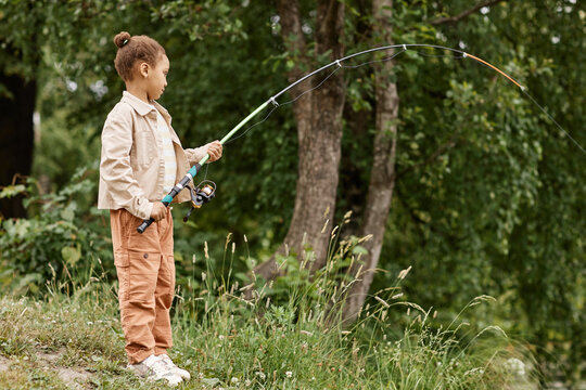 Full length side view portrait of black little girl fishing by river in nature with fishing rod bending, copy space 