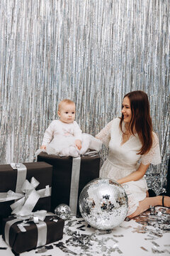 foil rain curtain for the photo zone. beautiful girl in a white dress and photo booth in silver style. mirror disco ball and silver confetti. mother and little daughter hugging
