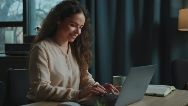 Portrait of joyful woman sitting in comfortable chair in front of laptop, chatting with boyfriend. Smiling girl wearing casual outfit looking at screen of computer and typing on keyboard. Indoors