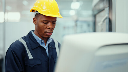 Portrait of African production engineer in safety wear operating CNC machine in the factory. A male factory worker concentrate on programming industrial machine to control the process of production.