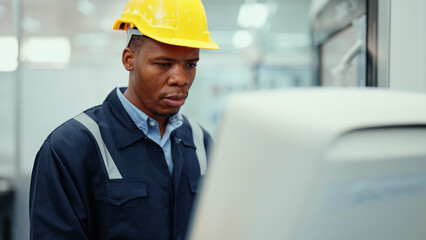 Portrait of African production engineer in safety wear operating CNC machine in the factory. A male factory worker concentrate on programming industrial machine to control the process of production.