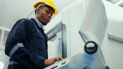 African production engineer in safety wear is operating CNC machine in the factory. A male factory worker with responsibility for programming industrial machine to control the process of production.