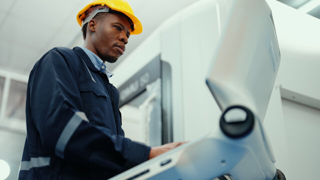 African production engineer in safety wear is operating CNC machine in the factory. A male factory worker with responsibility for programming industrial machine to control the process of production.