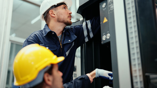 Two Caucasian production engineers in safety wear are assisting in adjusting and maintaining CNC machine in the factory. Male factory workers are examining the industrial machine to find an error.