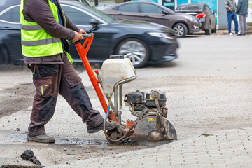 A road worker cuts with a petrol cutter and a diamond wheel the old asphalt on the carriageway of a city street. - obrazy, fototapety, plakaty