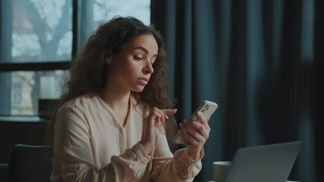 Young Caucasian woman sitting at table with laptop, holding smartphone in hands and posting on social media. Footage of focused girl in elegant blouse typing message in device. Indoors