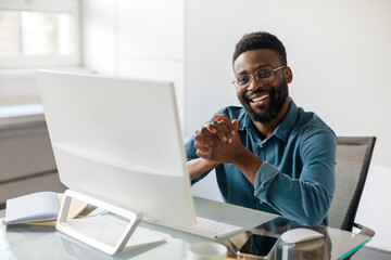 Positive african american male entrepreneur sitting at workdesk in office in front of computer and smiling at camera
