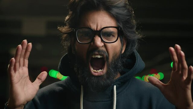 Male portrait at dark parking lot irritated emotional angry Indian bearded man in glasses shouting screaming yelling with mad anger aggressive Arabian guy shout scream to camera with furious stress