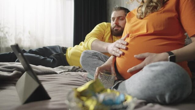Happy man touching pregnant woman's belly. Caucasian couple in love who are expecting a baby are looking at a tablet together.