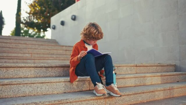 Back to school. A cute child with a backpack sits on the stairs near the school and reads a textbook. Elementary school student studying homework after class.