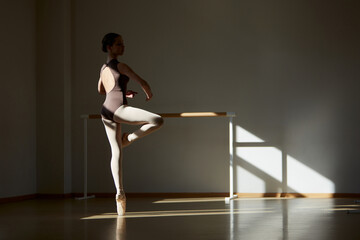 Beautiful teen girl, ballet dancer training, dancing on pointee at dance school with ballet barre on daytime with sunlight. Concept of classical ballet, dance art, education, beauty, choreography