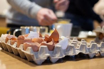 Broken egg shells in large carton box. Out of focus hands separating egg yolks in the background. Cooking with eggs.