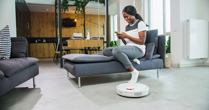 Smiling young african american lady house owner is sitting on sofa in living room using smartphone while robotic hoover is cleaning floor doing domestic work. Inventions and everyday life concept.