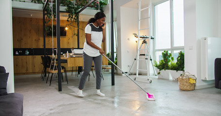 Beautiful african american woman mopping floor in living room with mop. Pretty housewife doing housework mopping while robotic vacuum cleaning floor. Female reading news browsing using smartphone.