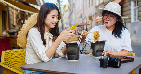 Portrait of happy Asian females sitting on restaurant terrace and eating chinese food outdoors and chatting. Young daughter having lunch with mother in cafeteria on street. Leisure concept