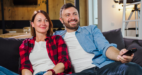 Obraz premium Portrait of young beautiful couple watching TV at home holding remote control, speaking, discussing, enjoying film together. Leisure activities while lockdown, young couple lifestyle concept.
