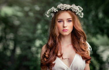 Beautiful red haired girl in white vintage dress and wreath of flowers standing under the tree and looking at camera. Fairytale story. Warm art work.