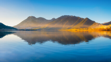 Obraz premium Lake and mountains in a valley at dawn. Reflections on the surface of the lake. Mountain landscape at sunrise. Foggy morning. Natural landscape with bright sunshine.