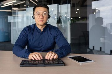 Portrait of serious successful specialist inside office, asian man with headset phone and keyboard sitting at desk and looking at camera, male online customer service call center worker.