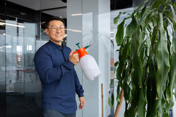 Portrait of successful smiling asian man inside office, boss watering flowers spraying flower pot...