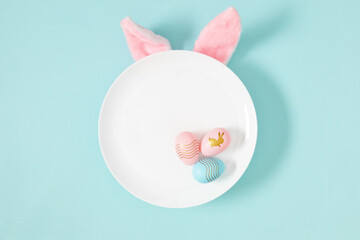 Easter holiday composition. Empty white plate with pink ears bunny and easter eggs on isolated pastel blue background. Easter concept. Flat lay, top view, copy space 