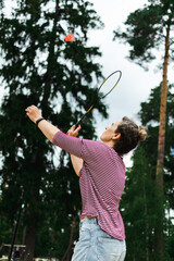 Outdoors against the forest and the sky on a cloudy cool day, a woman plays badminton, hits a red shuttlecock with a racket, copy space.